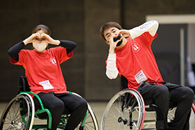 写真：車いすに乗りハートマークを手で作っている2人の学生