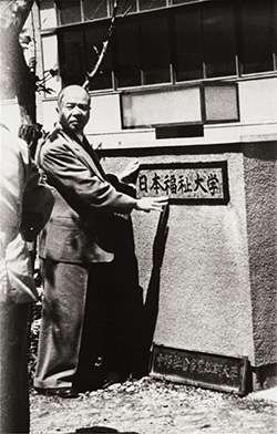 写真：日本福祉大学の銘板前に立つ鈴木修学先生のモノクロ写真。