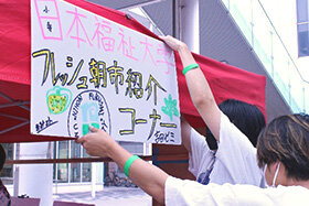 写真：「日本福祉大学 フレッシュ朝市紹介コーナー」と書いたパネルを設置する学生